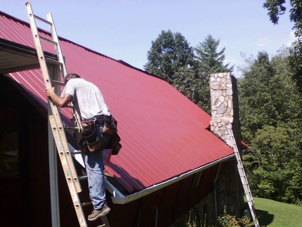 Roofer on a ladder beside a red metal roof and stone chimney — Soffit & Fascia Repair by Roofing Brothers in Morganton